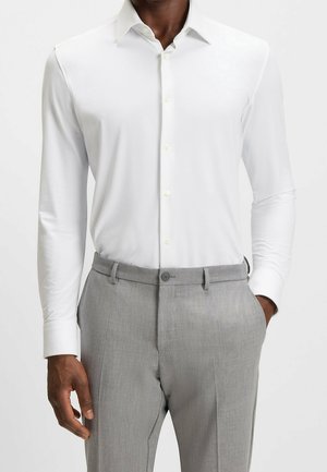 Homme portant une chemise blanche ajustée à manches longues rentrée dans un pantalon gris clair sur mesure, les mains dans les poches.