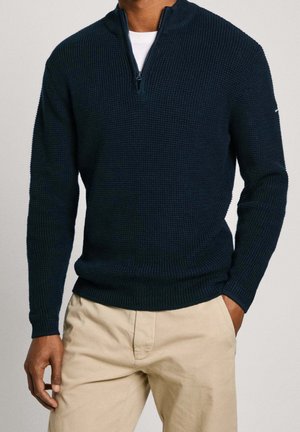Pull en maille bleu marine avec un design à zip quart, tissu texturé, et poignets et ourlet côtelés. Associé à un pantalon de couleur claire.