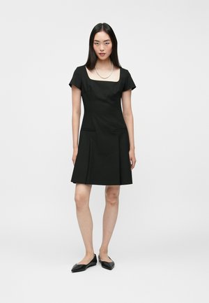 Black fitted dress with a square neckline, short sleeves, and two side pockets. Smooth fabric with a flared hem, paired with black flats.