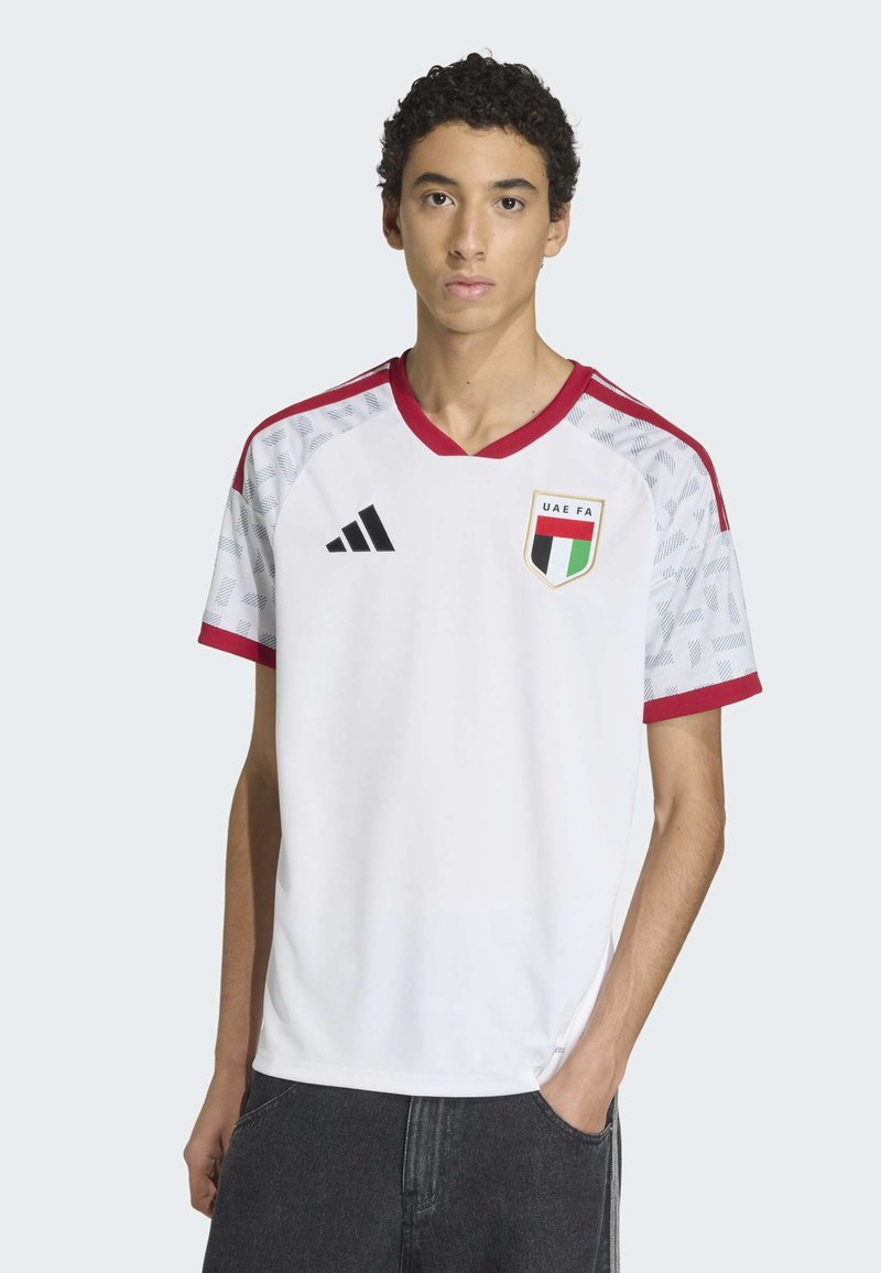 Maillot de football blanc avec des accents rouges et des manches à motifs. Présente un col en V, le logo adidas et le badge de la Fédération de football des Émirats arabes unis sur la poitrine.