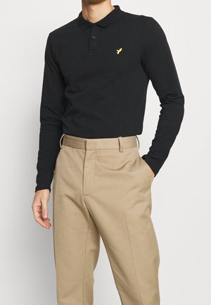 Homme portant un polo noir à manches longues avec un logo d’oiseau jaune, rentré dans un pantalon beige taille haute, debout avec une main dans la poche.