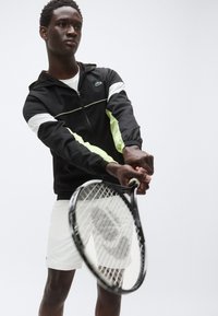 Črni športni suknjič z belimi in svetlo zelenimi poudarki, z zadrgo in kapuco; nošen čez bele kratke hlače, medtem ko drži teniški lopar.