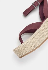 Burgundy mockaskesandal med vävd juteplattformssula, utrustad med en spännesträng och korsdesign, som framhäver textur och hantverk.