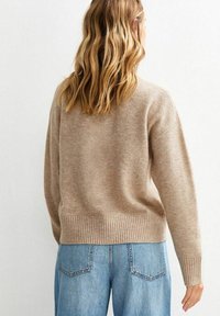 Pull en maille beige avec un ourlet et des poignets côtelés, présentant une coupe décontractée. Porté par-dessus un jean bleu clair. Texture douce visible.