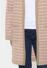 Beige kardi med horisontella rosa ränder, öppen front, två framfickor, ribbad ärm och textilstruktur. Bärs över en vit topp och mörka jeans.