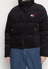 Černá žebrovaná prošívaná bunda s vysokým límcem, patentními knoflíky, kapsami na zip po stranách a malou značkovou nášivkou na hrudi.