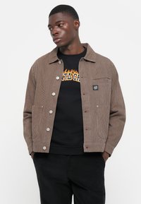 Bruine gestreepte jas met zilveren knopen en een borstzak, gedragen over een zwart sweatshirt. Model poseert tegen een effen achtergrond.