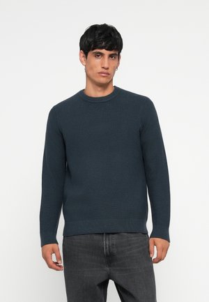 Dark blue ribbed sweater with a round neckline and long sleeves, made from textured knit fabric. Paired with gray jeans.