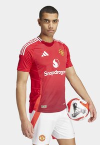 Maillot rouge de Manchester United avec un short blanc ; les caractéristiques incluent des épaules rayées, un logo et un texte de parrainage. Tient un ballon de football à motifs.