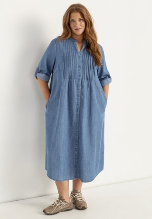 Frau mit langen roten Haaren, die ein blaues, geknöpftes Denim-Midikleid mit hochgekrempelten Ärmeln und beigen Sneakers trägt, vor weißem Hintergrund stehend.