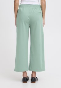 Lichtgroene wijde broek van zachte stof, met een elastische tailleband en een enkele achterzak. Er worden platte zwarte schoenen gedragen.