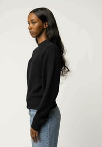 Schwarzer gerippter Pullover mit hohem Kragen, langen Ärmeln und strukturiertem Stoff, kombiniert mit hellblauen Jeans, seitlich betrachtet.