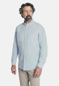 Camisa azul claro de botones con mangas largas, tejido ligeramente texturizado y cuello clásico, combinada con pantalones beige claros.