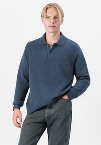 Sudadera de polar azul con cuello, que presenta una superficie texturizada, tres botones y mangas largas enrolladas, combinada con jeans de mezclilla gris.