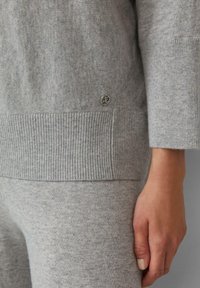 Nahaufnahme einer Person, die einen passenden hellgrauen Strickpullover und eine Hose trägt. Man sieht die Hand und ein kleines Metall-Logo am Saum des Pullovers.