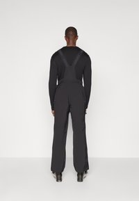 Schwarze Latzhosen in voller Länge mit verstellbaren Trägern, die über eine glatte Stofftextur und Reißverschlusstaschen verfügen. Eignen sich für verschiedene Aktivitäten.