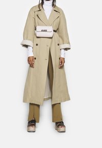 Beige linned trenchcoat med store klapper og opredte ærmer, hvid turtleneck, brune udvidede bukser og sorte platform sandaler.