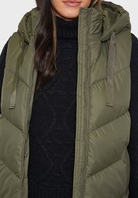Gilet en doudoune vert olive avec une capuche doublée, fermeture éclair et cordons de capuche réglables, superposé sur un pull noir texturé.