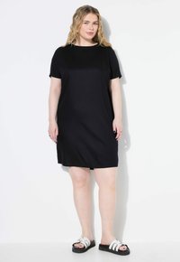 Schwarzes Kurzarmkleid mit lockerem Schnitt, aus weichem Stoff, der knapp über dem Knie endet; kombiniert mit weißen Sandalen mit auffälligen schwarzen Sohlen.