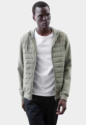 Młody mężczyzna ubrany w jasnobiały, pikowany, zapinany na zamek kaptur na białej koszulce i czarnych spodniach, stojący na jednolitym tle.