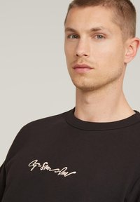 Black cotton sweatshirt with a crew neck, featuring a light-colored handwritten logo on the chest, smooth texture, and a relaxed fit.