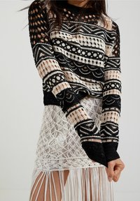Schwarzer und cremefarbener Strickpullover mit durchbrochenen Details und geometrischen Mustern, kombiniert mit einem weißen Makramee-Rock mit Fransen an den Rändern.
