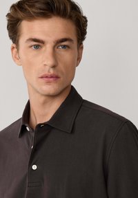 Polo shirt marrone scuro con colletto strutturato e tre bottoni. Texture liscia, maniche corte e un sottile dettaglio della tasca sul petto.