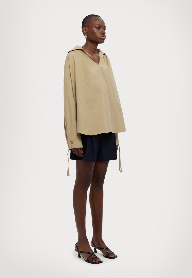 Maglione beige oversize con cappuccio a punta, maniche lunghe e polsini con cordoncino, abbinato a pantaloni corti blu navy e scarpe con tacco nere a strisce.