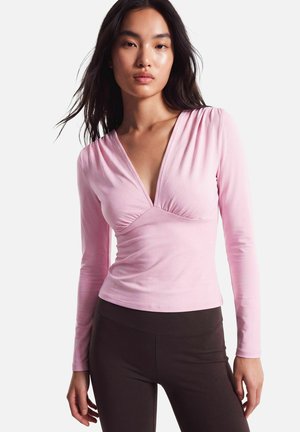 Femme aux longs cheveux foncés portant un haut rose clair à manches longues avec col en V et un pantalon foncé, debout devant un fond clair uni.