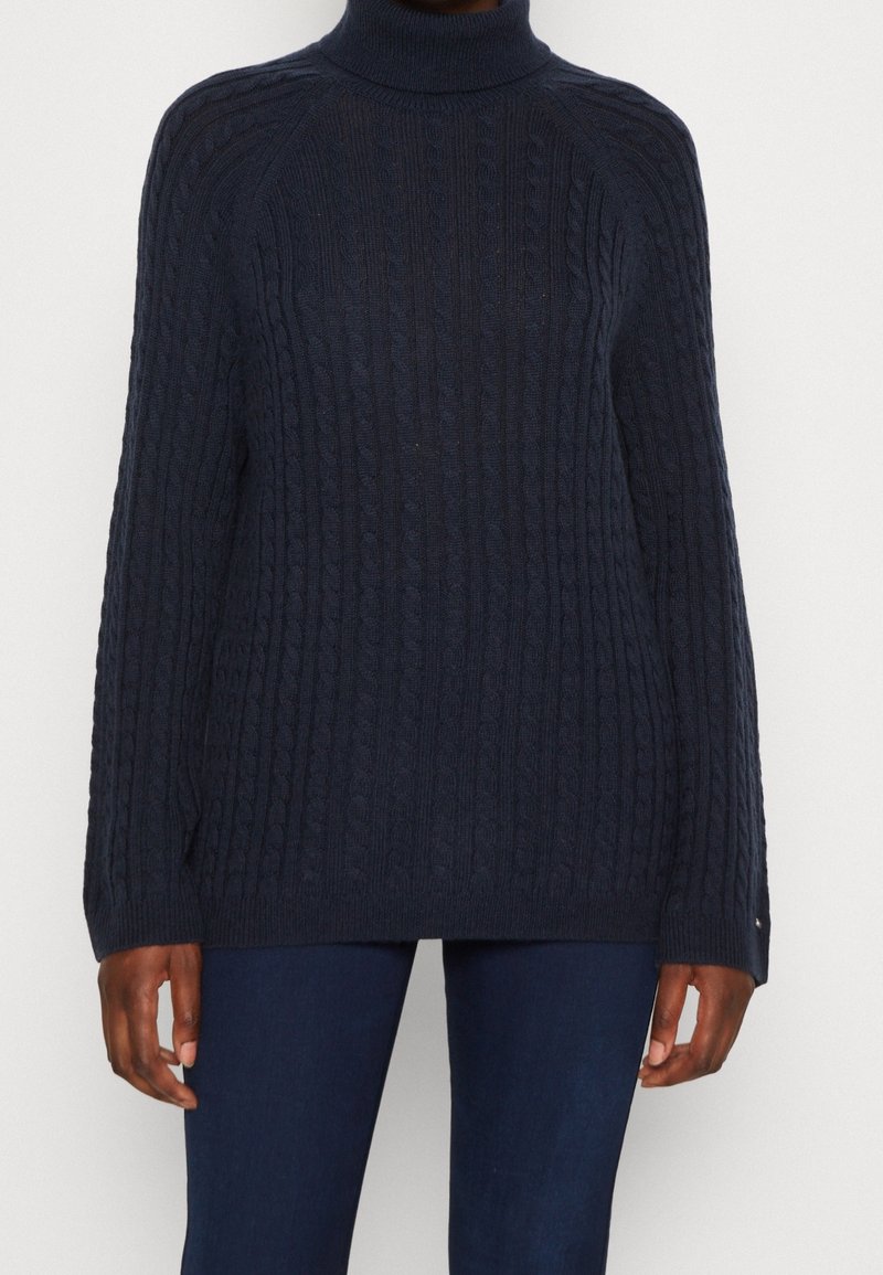 Pull à col roulé bleu marine foncé avec motif torsadé, porté avec un pantalon bleu foncé, montré des épaules aux hanches sur une personne à la peau brun moyen foncé.