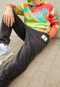 Kapuze mit mehrfarbigem Batikmuster, schwarze ausgefranste Sweatpants mit Logo-Patch und graue Sneakers mit weißen Akzenten.