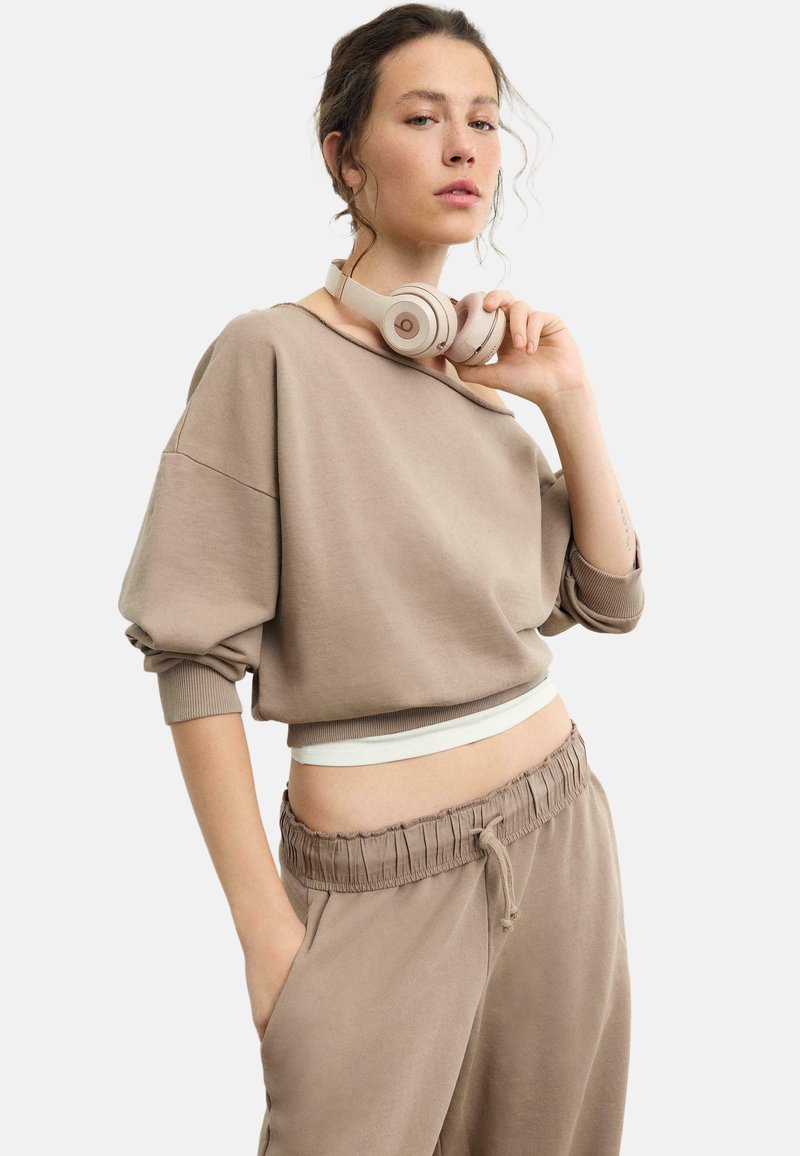 Ung kvinde iført beige off-shoulder sweatshirt og matchende sweatpants, med beige over-ear hovedtelefoner om halsen.