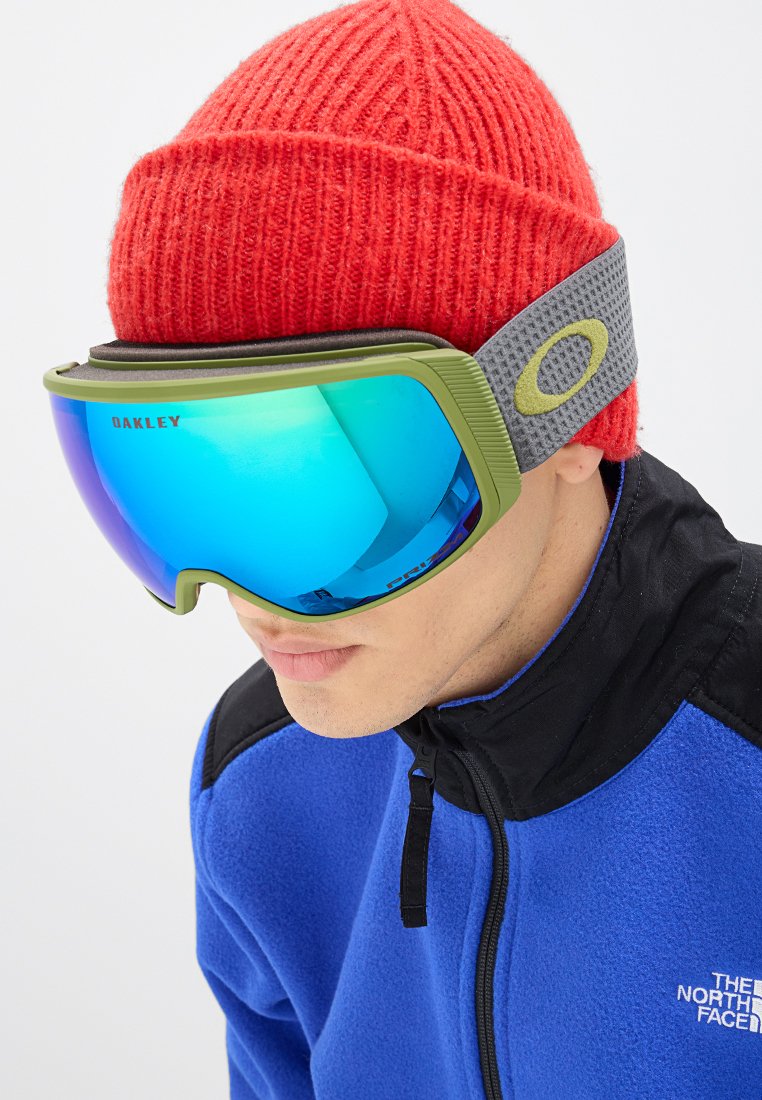 Zielona rama gogli narciarskich z niebieskimi lustrzanymi soczewkami, fakturowany szary pasek, noszone z czerwonym dzianinowym czapkiem i niebieską polarek.
