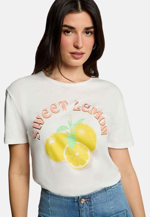 Femme portant un t-shirt blanc avec un motif de citrons jaunes et le texte "Sweet Lemon", associé à un jean bleu taille haute.