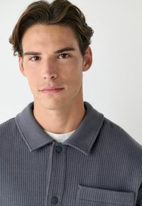 Grau gerippte Shirtsjacke mit Kragen, Knopfverschluss und einer Brusttasche. Verfügt über einen strukturierten Stoff und eine lockere Passform.