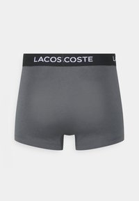 Grijze boxershorts met een zwarte tailleband waaraan witte "LACOSTE" tekst is toegevoegd. De stof lijkt glad en rekbaar, met een aansluitend ontwerp.