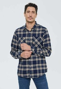 Chemise en flanelle pour homme avec un motif à carreaux bleu marine et beige, devant à boutons, deux poches poitrine et manches longues. Texture douce.