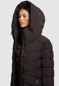 Schwarze Daunenjacke mit großer Kapuze, ausgestattet mit einem Reißverschluss und einem plüschigen, gesteppten Design. Strukturierter Stoff mit subtiler Glanzwirkung.