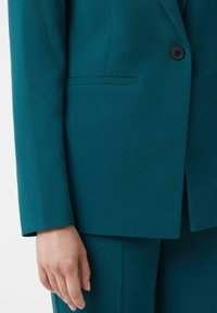 Zbliżenie na tealową marynarkę z pojedynczym czarnym guzikiem i lewą ręką osoby swobodnie opartą na boku.