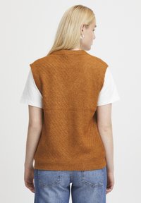Rostfarbener gestrickter Pullunder mit einem Zickzackmuster, kurzen Ärmeln und einem gerippten Saum, kombiniert mit einem weißen T-Shirt und blauen Jeans.