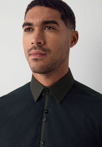 Camicia abbottonata verde scuro con colletto a trama e motivo. Presenta un tessuto liscio e cinque bottoni lungo la parte anteriore.