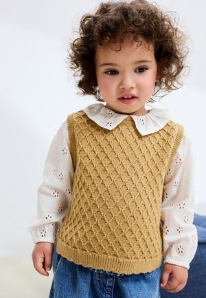 Gilet di maglia giallo senape con motivo a rombi, indossato sopra una camicetta bianca con dettagli a occhiello e colletto Peter Pan.