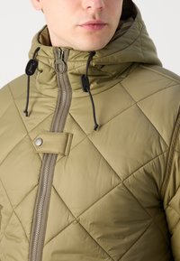 Veste khaki matelassée avec capuche, comportant une fermeture éclair intégrale, des cordons de serrage et des détails à bouton-pression. Tissu lisse et léger avec des coutures géométriques.