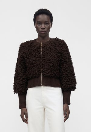 Woman wearing a dark brown textured zip-up sweater with puffed sleeves and white high-waisted pants, standing against a plain background.