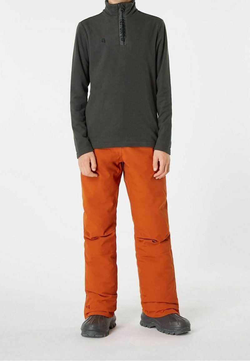 Personne portant un haut à manches longues gris foncé zippé, un pantalon isolant orange vif et des bottes d'extérieur noires, debout devant un fond uni.