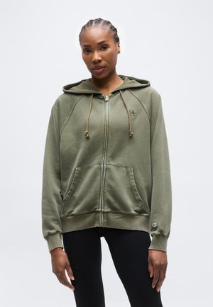 Γυναίκα με πλεξούδες που φοράει χακί φούτερ με φερμουάρ και μπροστινές τσέπες, καθώς και μαύρα κολάν, στέκεται μπροστά σε απλό φόντο.