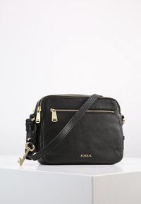 Schwarz strukturierten Leder Fossil Umhängetasche mit goldenen Reißverschlüssen, verstellbarem Tragegurt, Vordertasche und dekorativem Schlüsselanhänger auf weißer Oberfläche.