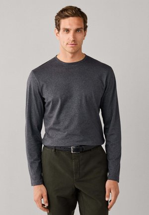 Mann trägt ein dunkelgraues Langarmshirt und olivgrüne Hose und steht vor einem einfarbigen hellgrauen Hintergrund.