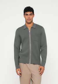 Grå strikket cardigan med glidelås foran, krage, lange ermer og brystlomme. Glatt tekstur med en avslappet passform.