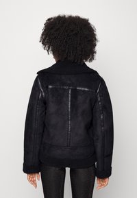 Chaqueta de ante sintético negra con cuello y puños de borreguito. Presenta costuras en contraste y una silueta amplia. Vista trasera; no se ven bolsillos.